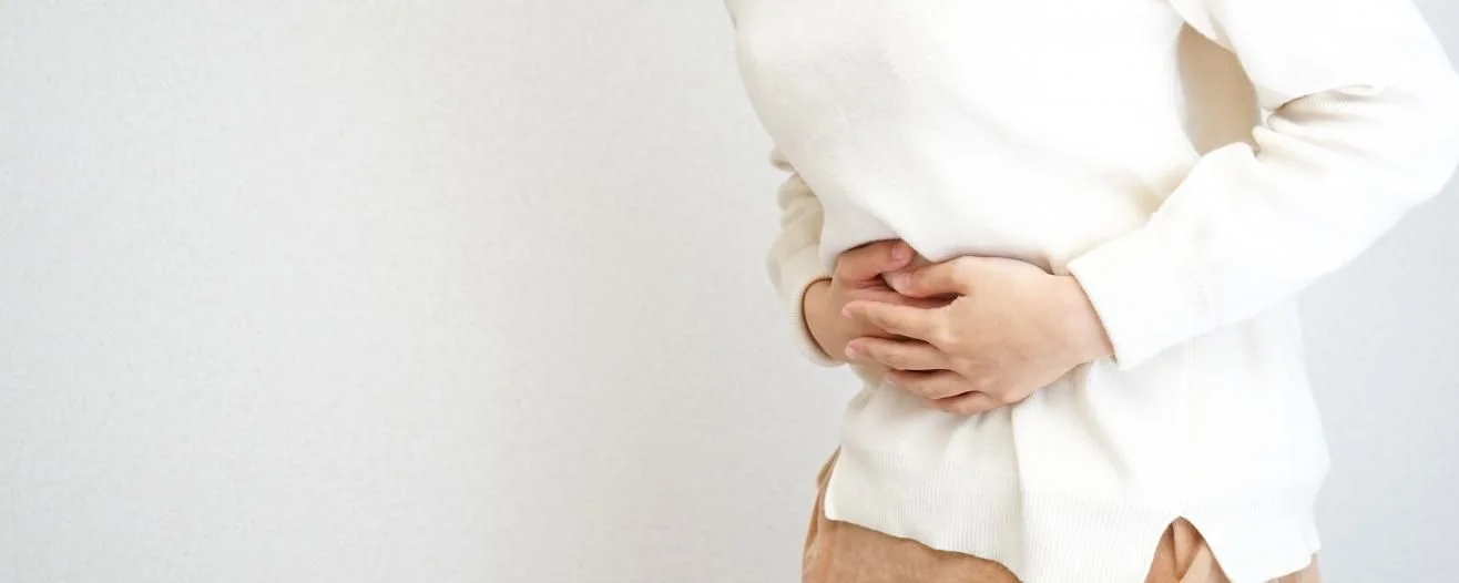A person wearing a white sweater holds their stomach, showing discomfort against a plain background