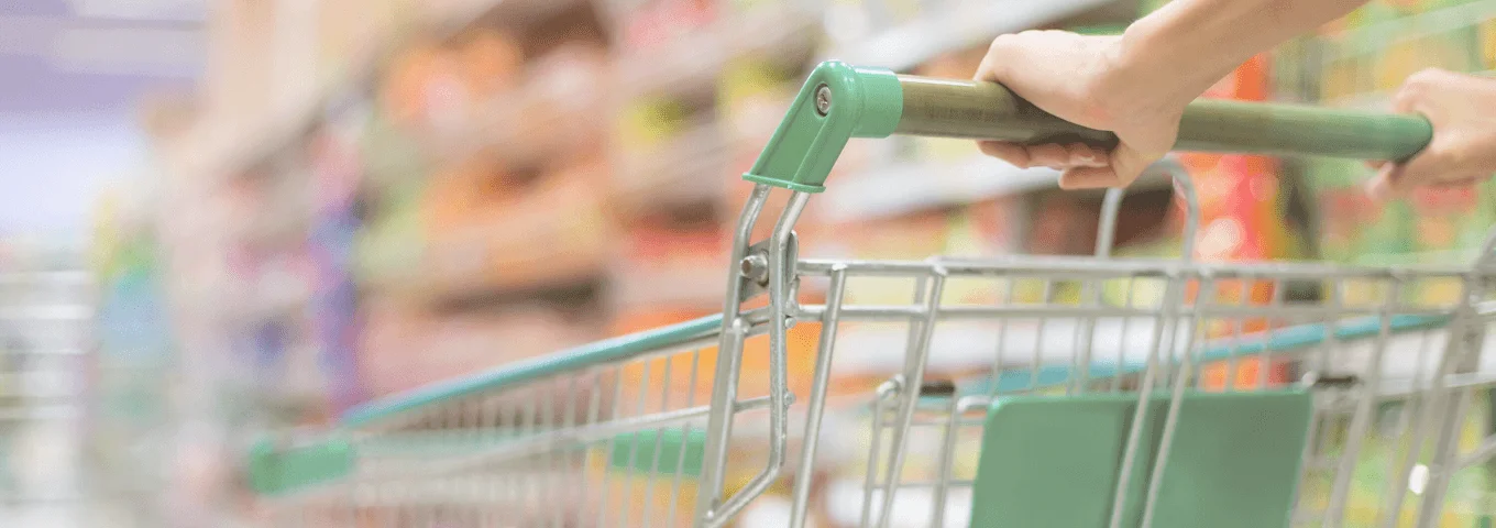 Eine Person schiebt einen Einkaufswagen durch einen Supermarktgang mit bunten Produkten im Hintergrund