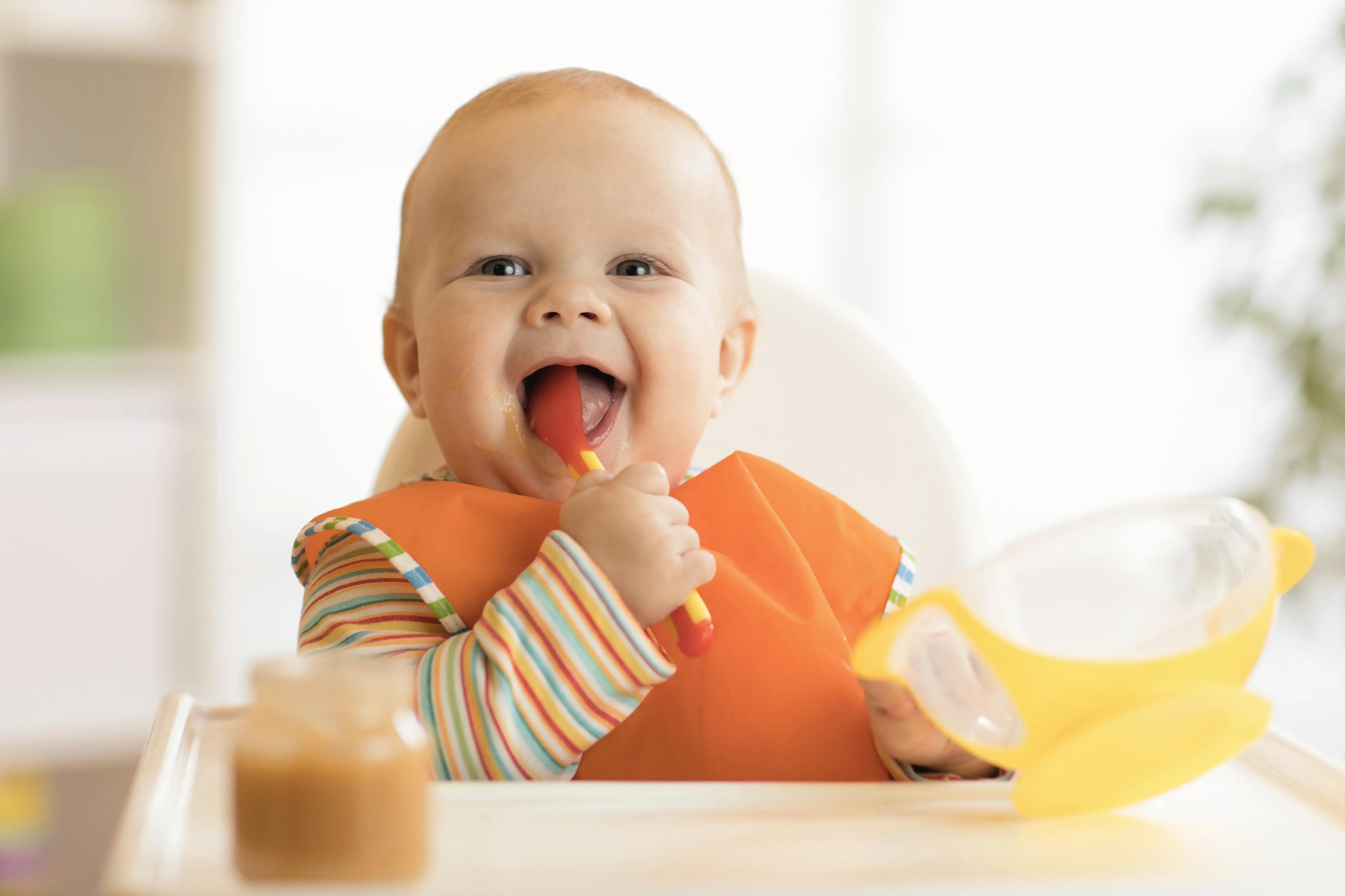 Bambino felice con un grande sorriso che tiene un cucchiaio e una ciotola gialla, in un ambiente luminoso