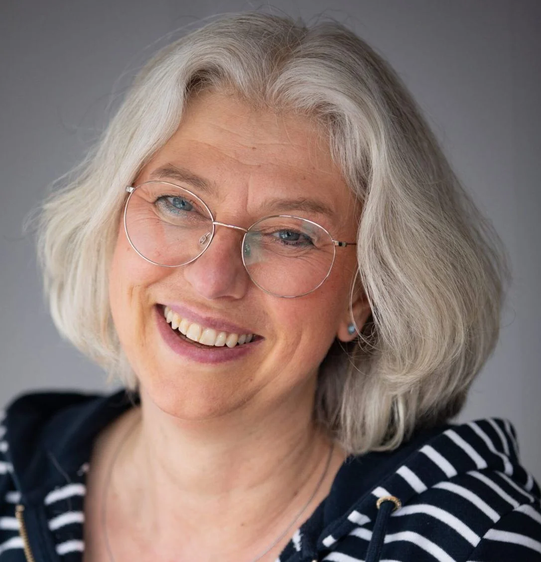 Smiling middle-aged woman with glasses and short gray hair, wearing a striped hoodie against a neutral background