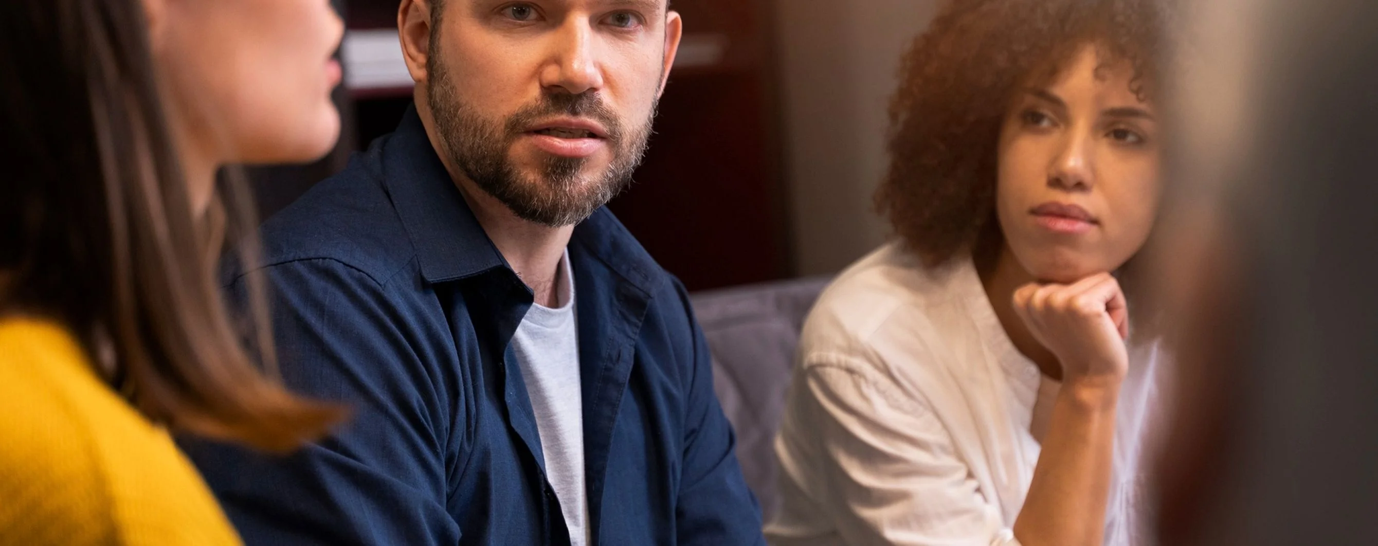 A man with a beard speaks attentively while two women listen in a modern meeting room