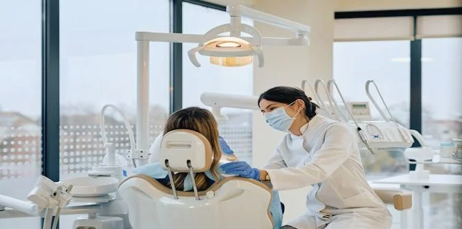 A dentist in a white coat and mask examines a patient seated in a dental chair in a modern clinic