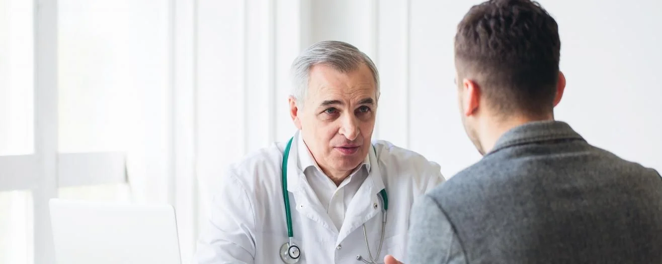 A doctor in a white coat with a stethoscope speaks with a male patient in a well-lit office