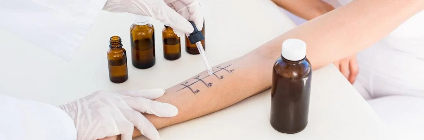 A person with gloved hands applies a dropper to marked skin on an arm, with several brown bottles nearby