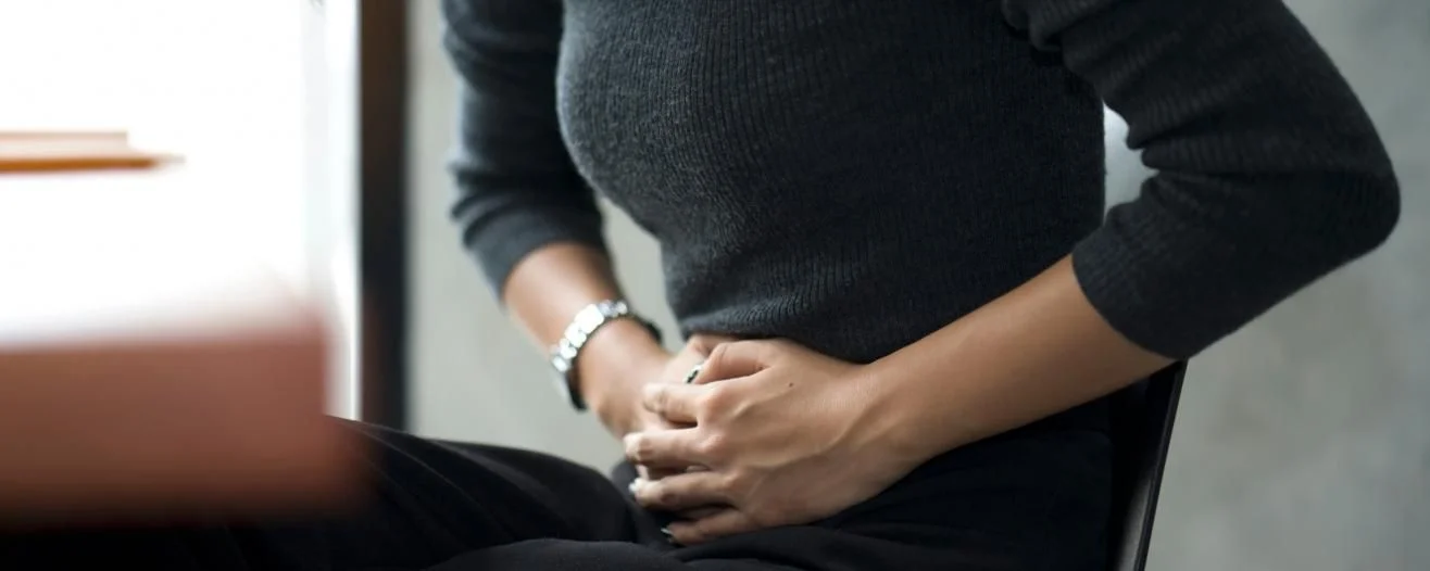 A person in a dark sweater is sitting, holding their abdomen with a pained expression, in an indoor setting