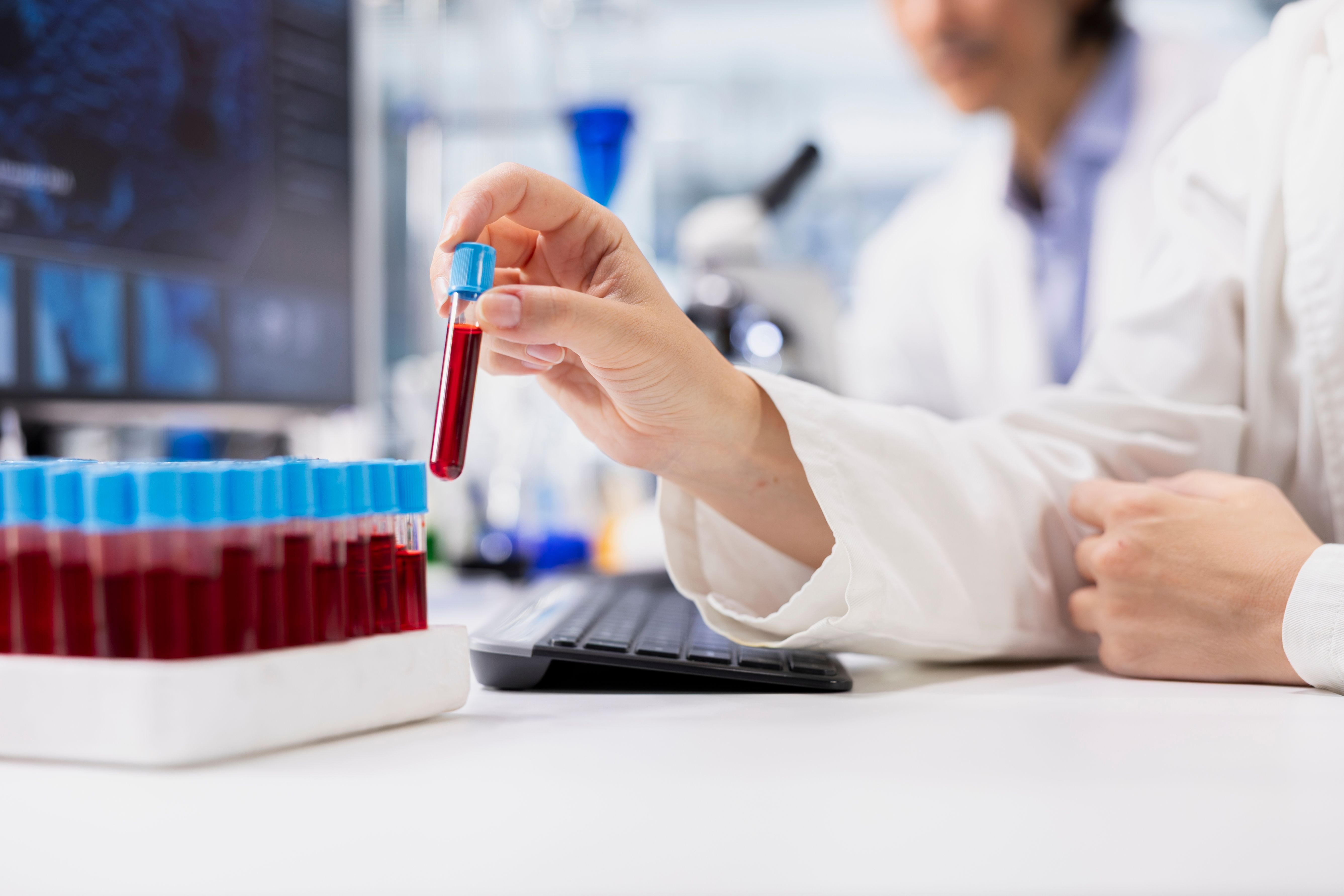 A researcher in a lab holds a test tube filled with red liquid, next to a rack of similar tubes and a computer keyboard