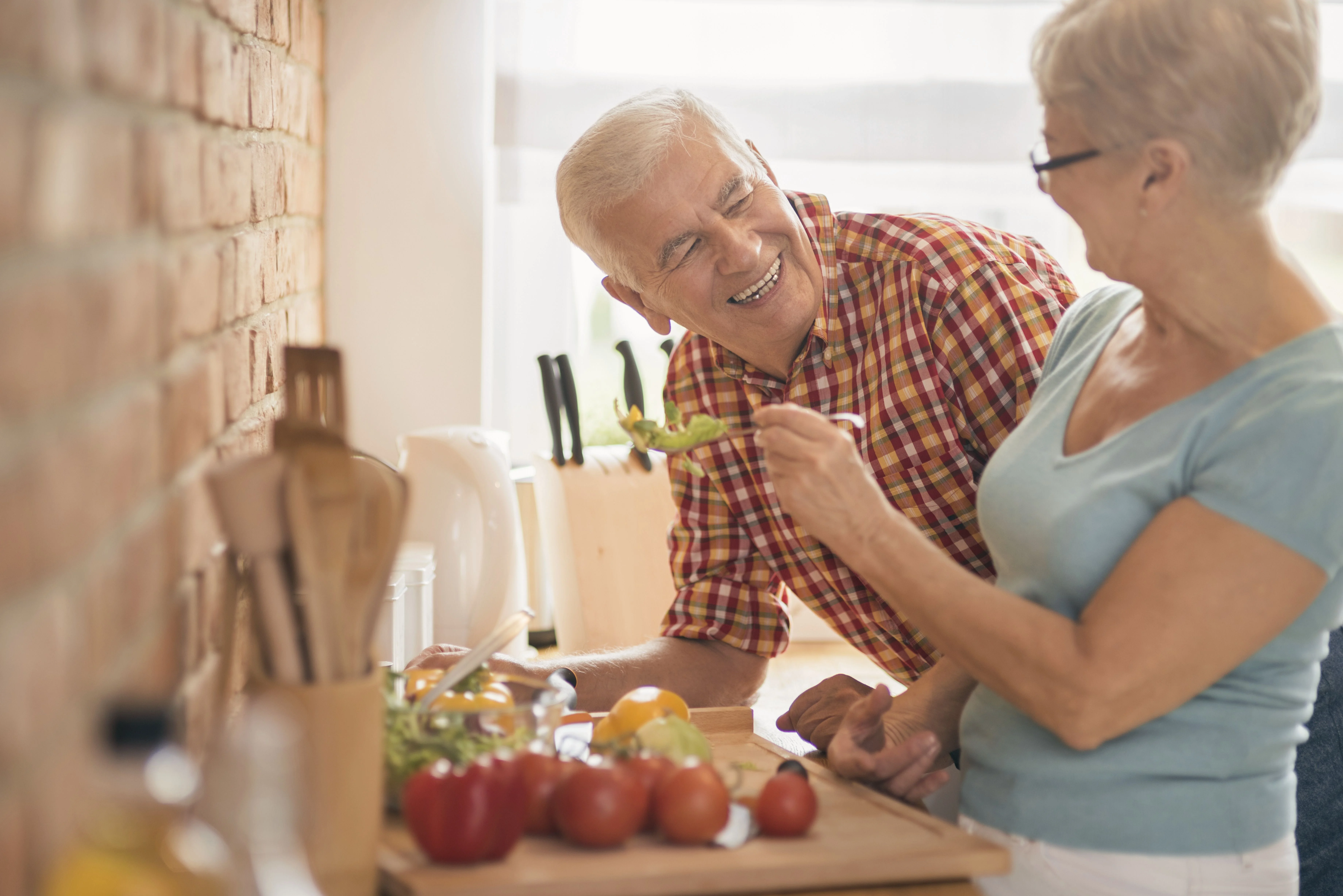 Un hombre mayor sonríe mientras disfruta de una comida con una mujer en una cocina acogedora llena de verduras frescas