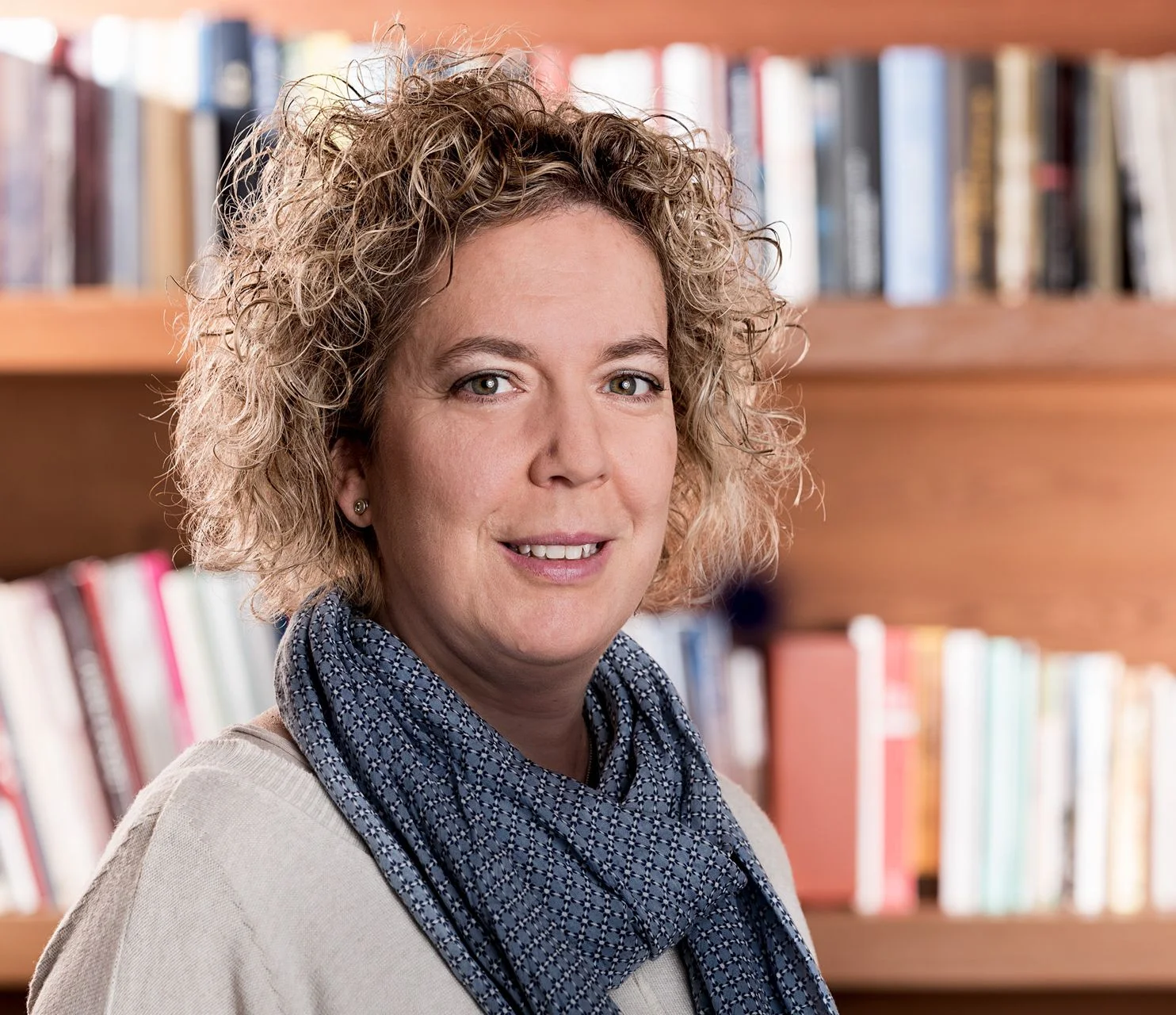Donna con capelli ricci sorride, indossa una sciarpa blu davanti a una libreria in legno piena di libri