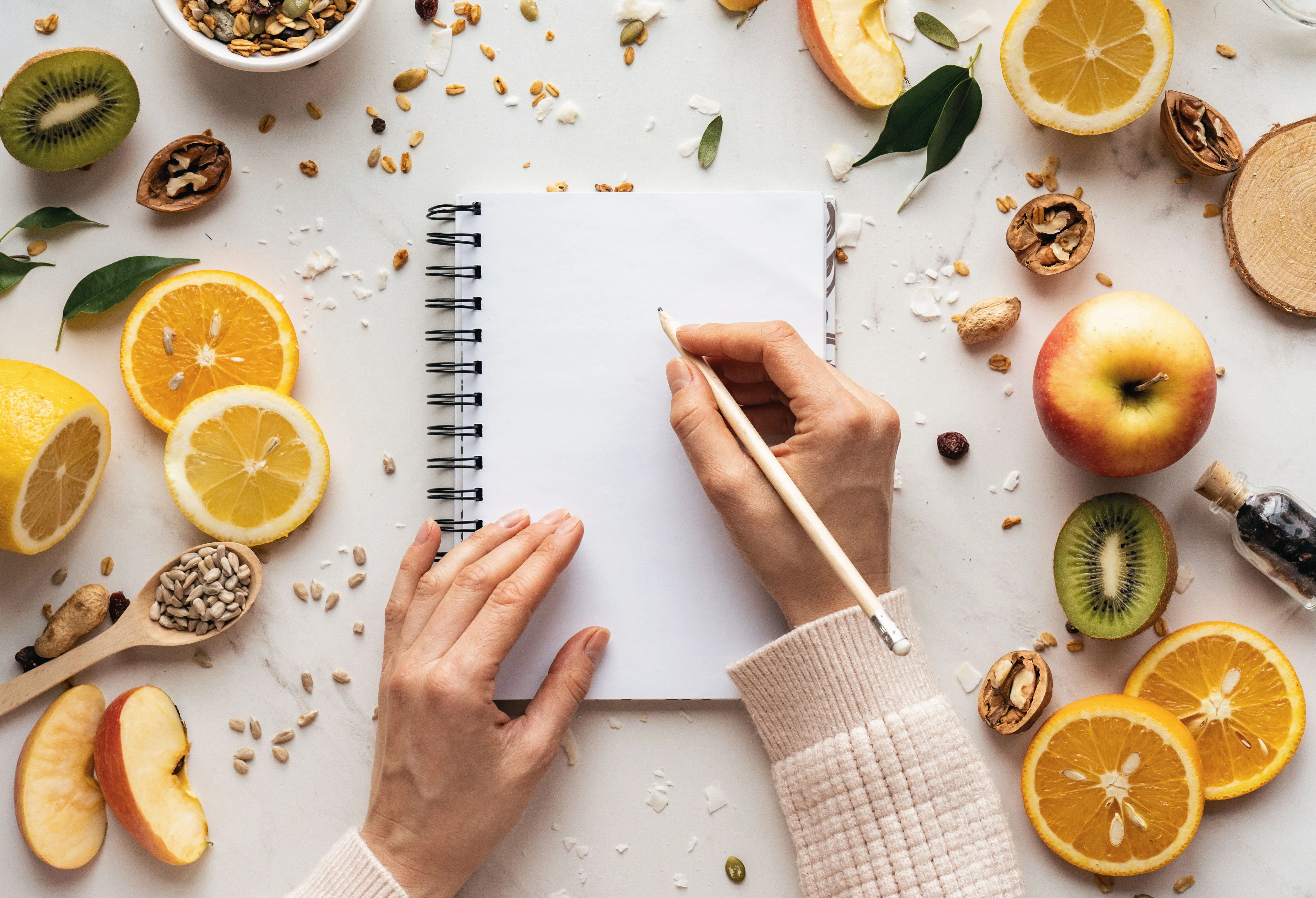 Una mano tiene una matita sopra un quaderno vuoto circondato da frutta, semi e noci su una superficie bianca
