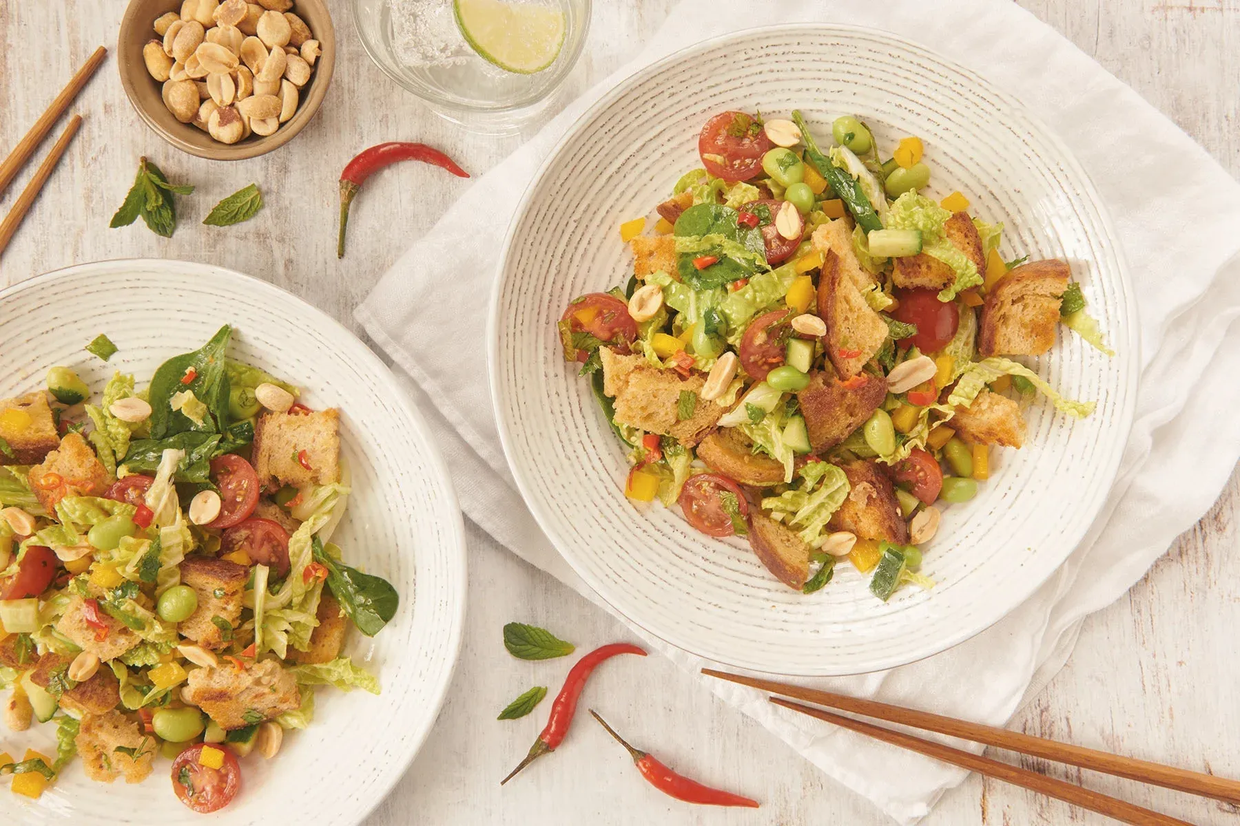 Dos tazones de ensalada colorida con tomates, verduras y pan crujiente, rodeados de cacahuetes y un chile