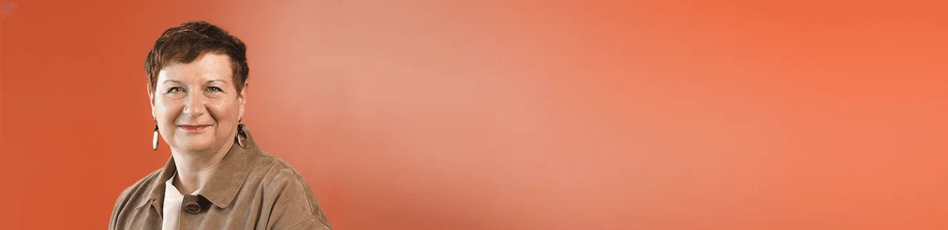 Una donna dai capelli corti sorride davanti a uno sfondo arancione caldo, indossando una giacca beige e orecchini