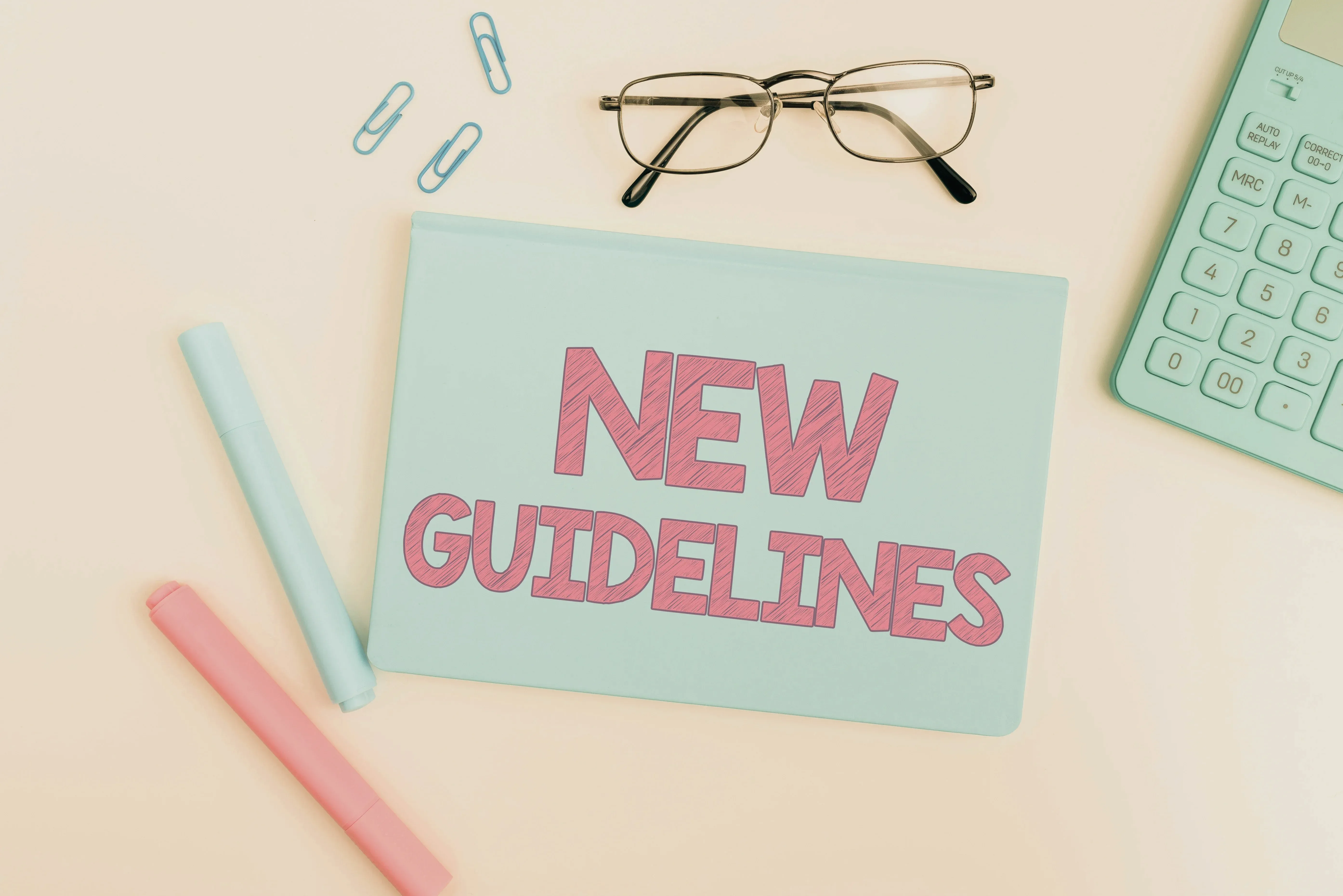 A pastel desk with 'NEW GUIDELINES' written on a blue notepad, two markers, glasses, and paper clips
