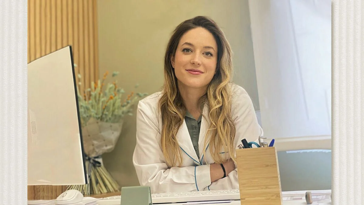 Una joven doctora con bata blanca sonríe a la cámara. Su oficina cuenta con una computadora y un arreglo floral
