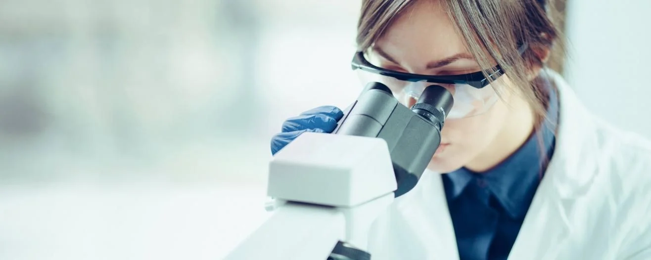 A focused researcher examines a specimen under a microscope, wearing safety glasses and a lab coat