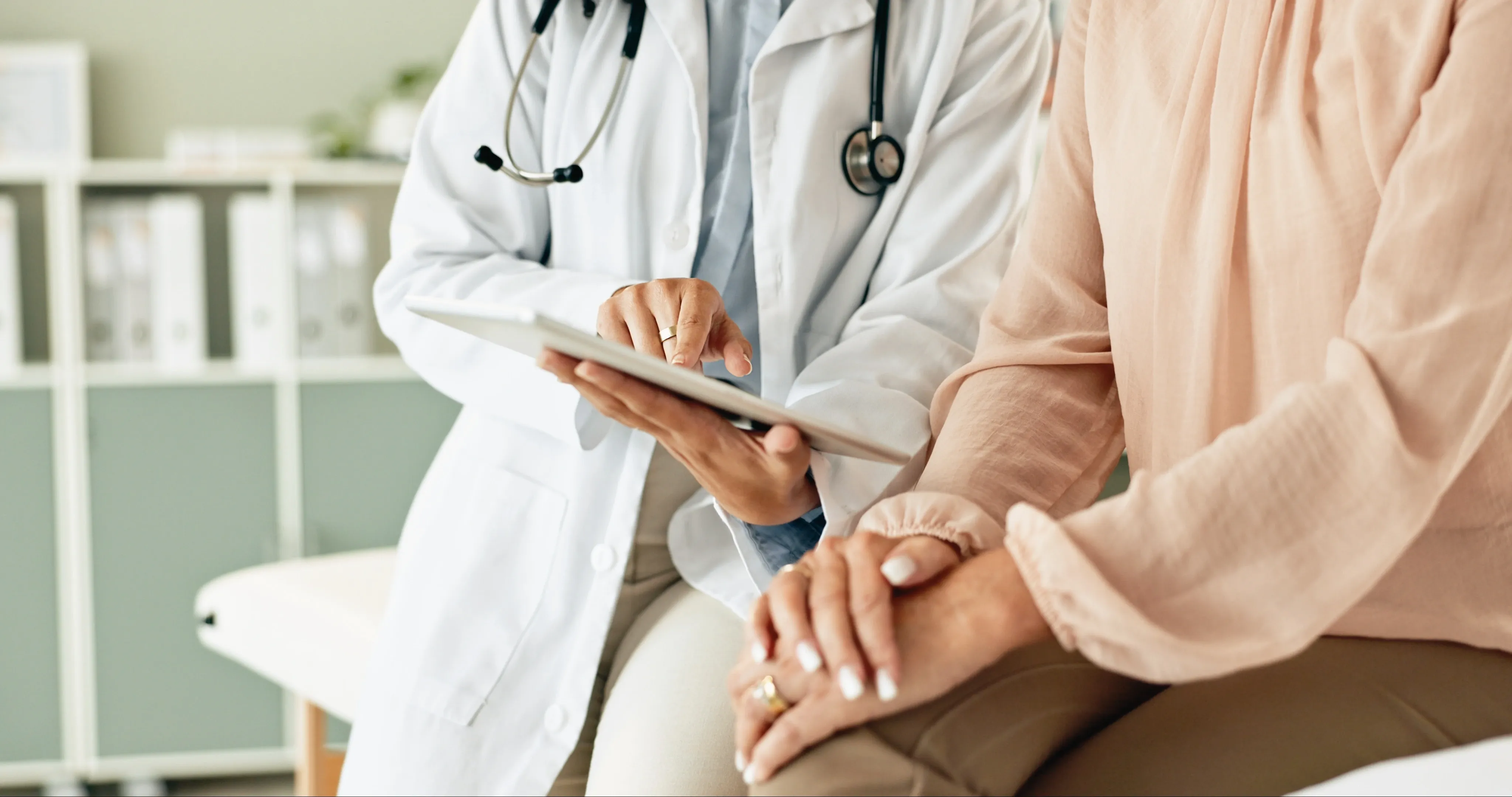 A doctor in a white coat examines a tablet while a patient rests hands on her lap in a medical office