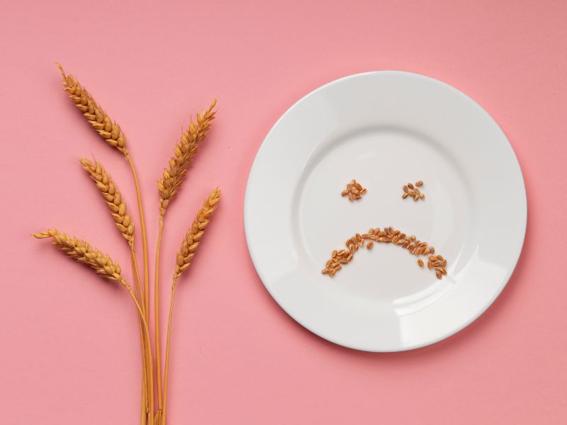 Wheat stalks beside a white plate featuring sad face made from seeds, set against a pink background