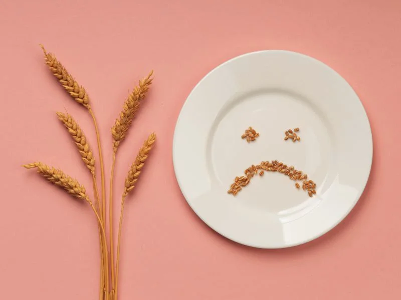 Un piatto bianco su uno sfondo rosa, con cereali disposti a formare un volto triste e spighe di grano accanto