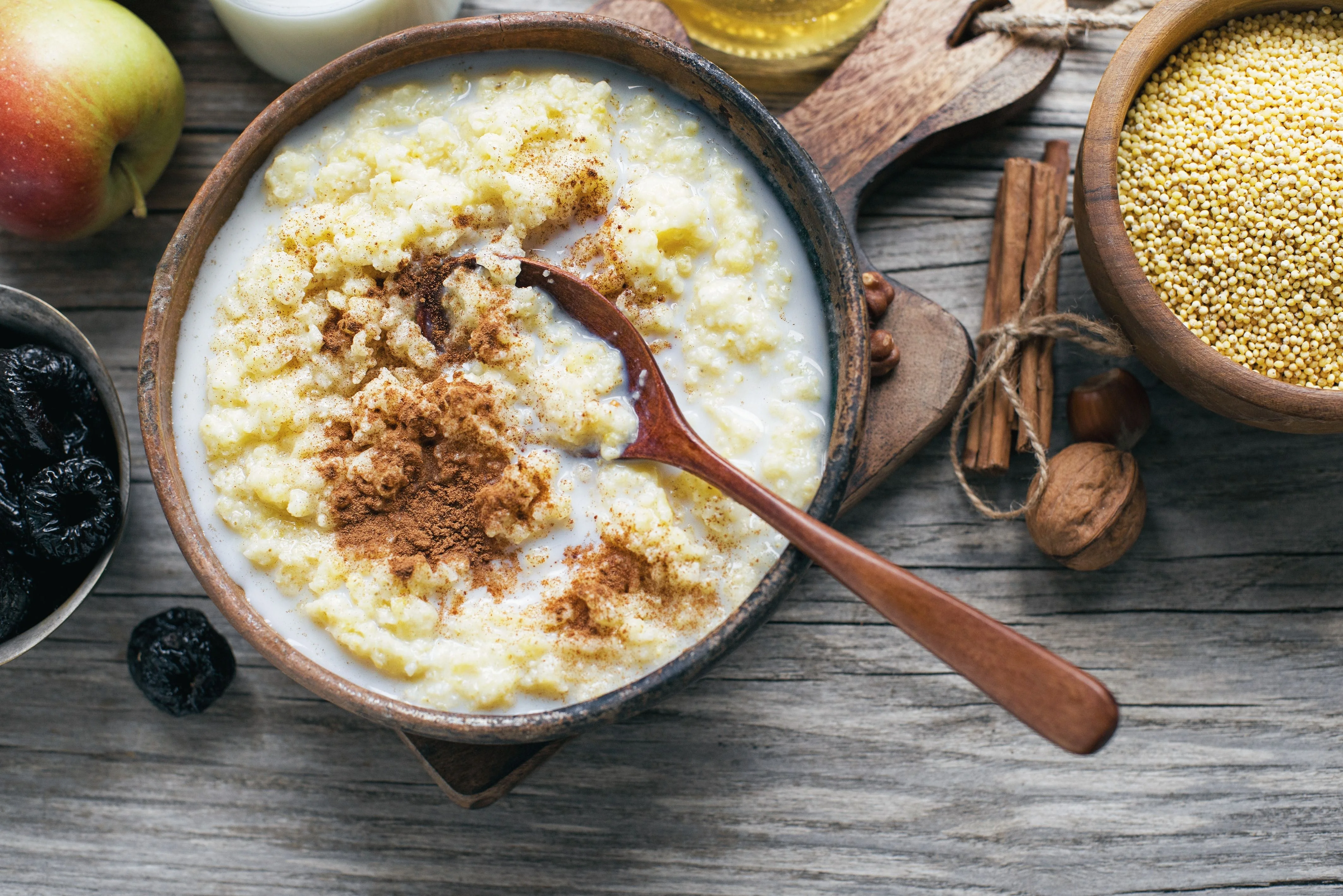 Un bol de crema de mijo con canela, rodeado de una manzana, ciruelas secas y especias