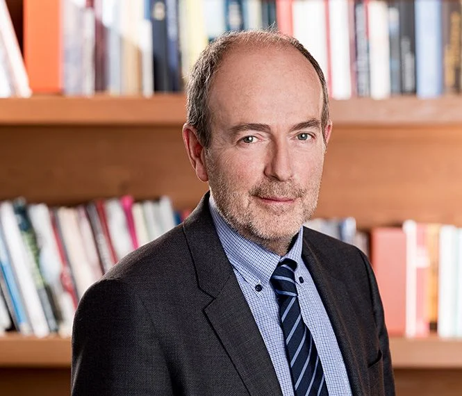 Uomo con un leggero sorriso, vestito con un compleanno e cravatta, si trova davanti a uno scaffale di libri