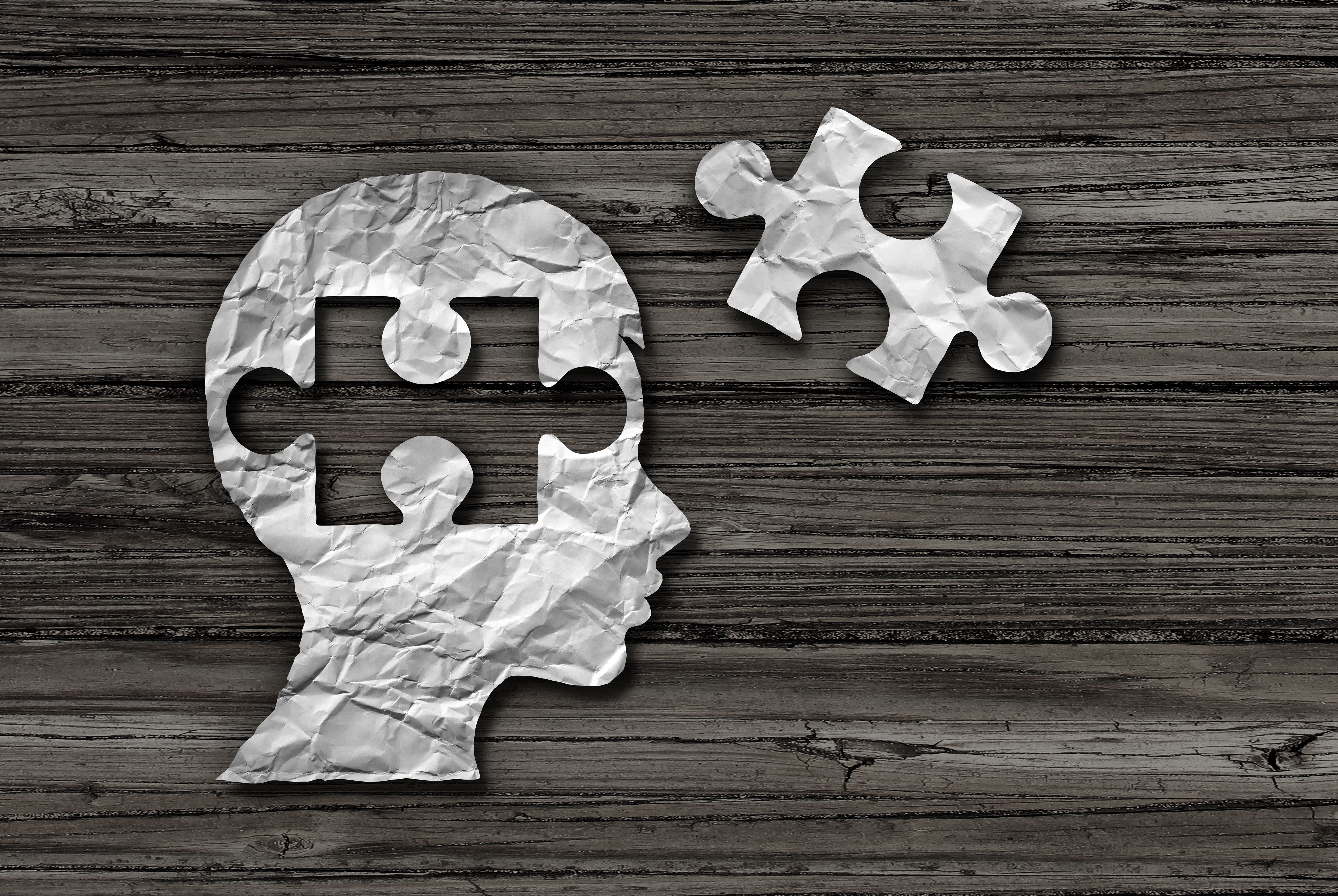 Crumpled paper cutout of a head with a puzzle piece missing, alongside a loose puzzle piece, on a wooden surface