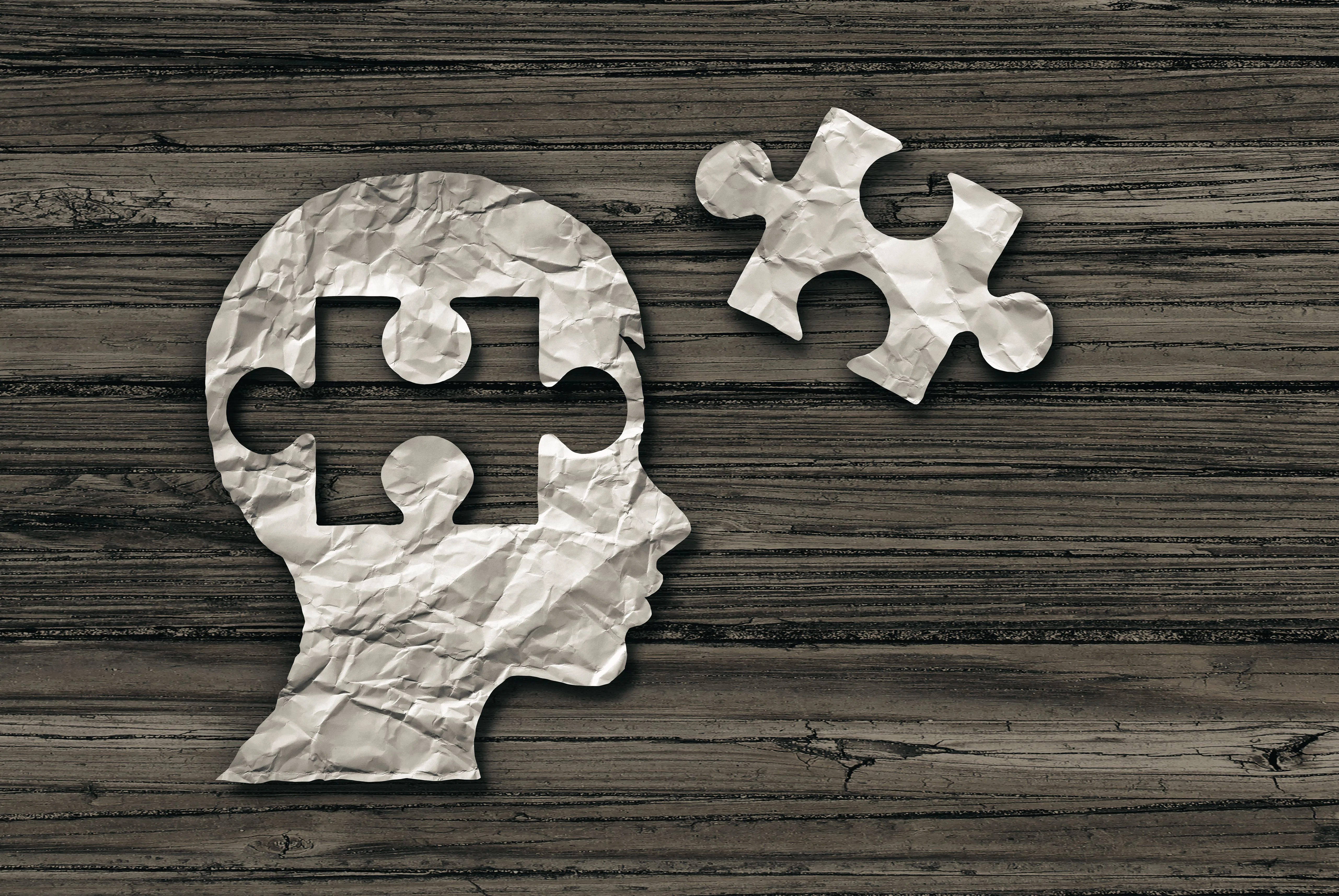 Crumpled paper cutout of a head with a puzzle piece missing, resting on a wooden surface