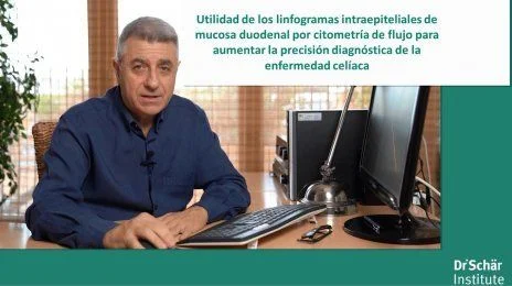 Un hombre se sienta en un escritorio con una computadora, hablando sobre métodos diagnósticos para la enfermedad celíaca, con luz del sol que entra por las persianas