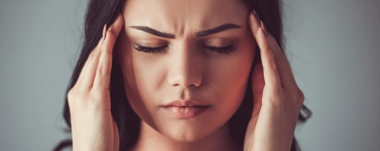 A woman with long dark hair grimaces, holding her head in pain, indicating a headache or discomfort