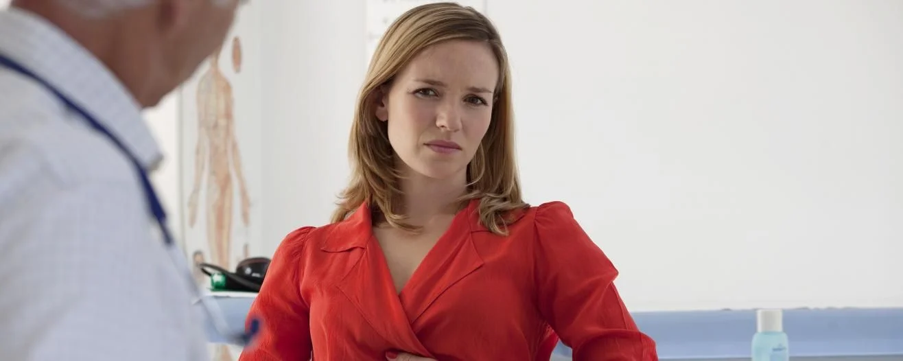 A woman in a red blouse looks concerned while speaking with a doctor in a medical office