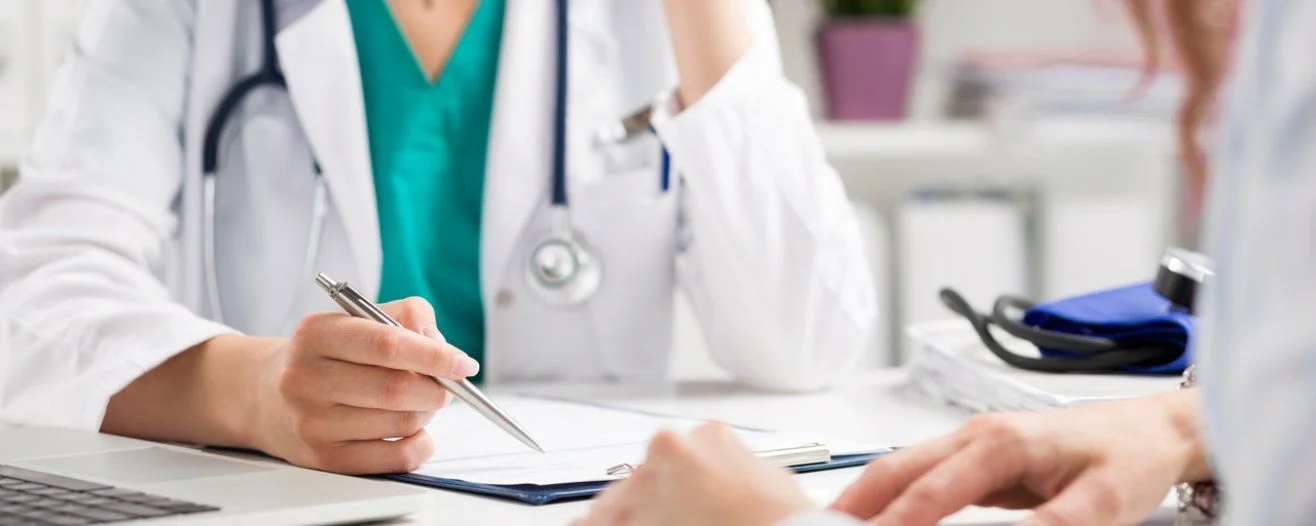 A doctor in a white coat reviews a document while taking notes, with a stethoscope around their neck
