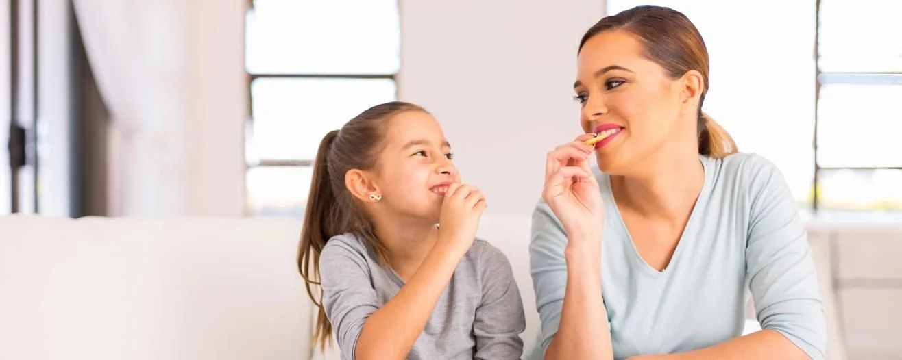 A mother and daughter share a joyful moment, eating snacks and smiling at each other in a bright living room