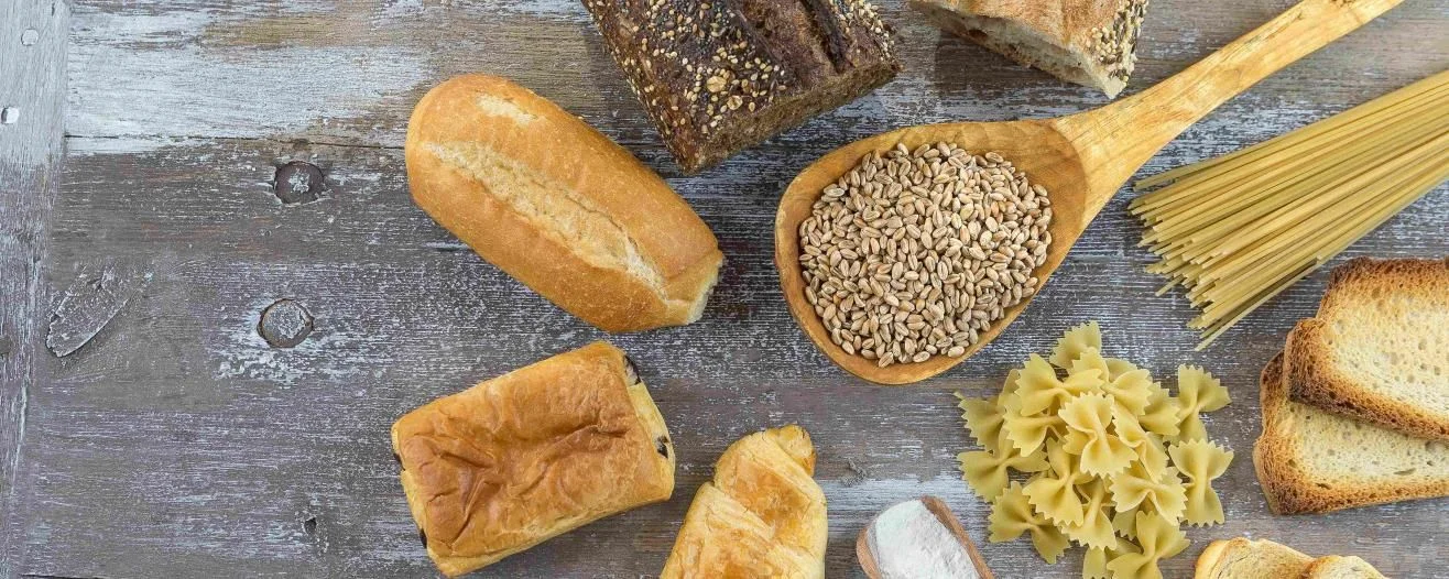 Una disposizione di vari tipi di pane, cereali, pasta e un cucchiaio di legno su una superficie in legno rustica