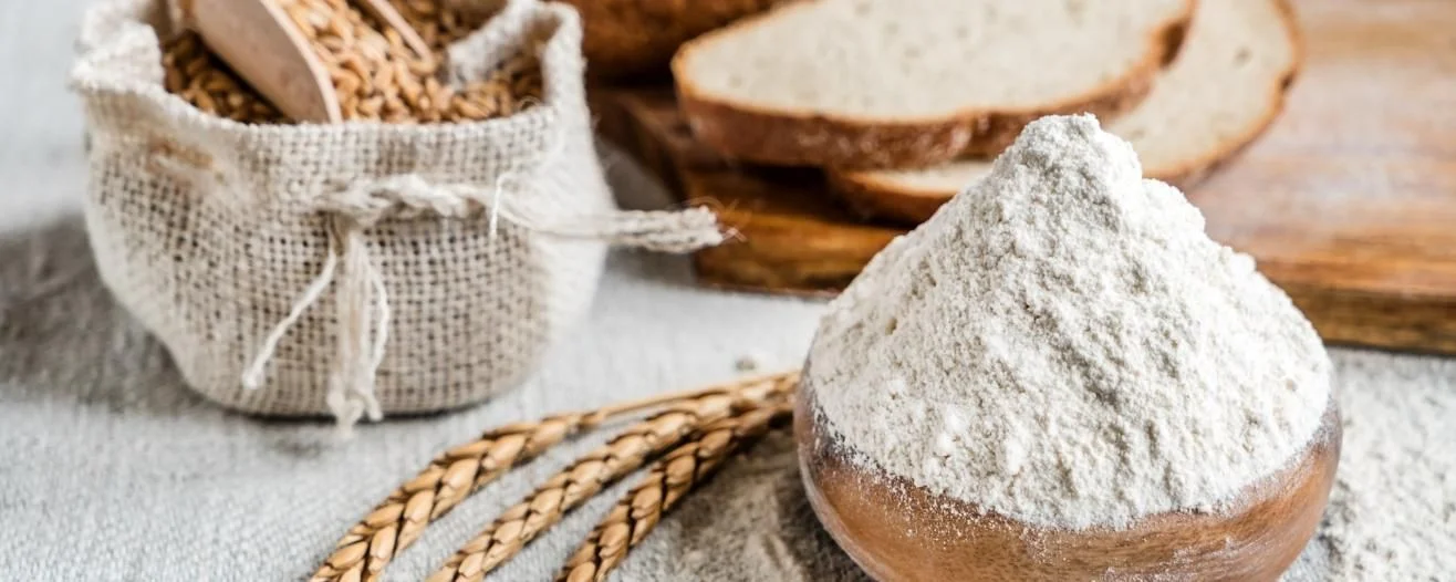 Brown bread slices, a wooden bowl of flour, a burlap sack with grains, and wheat stalks on a textured surface