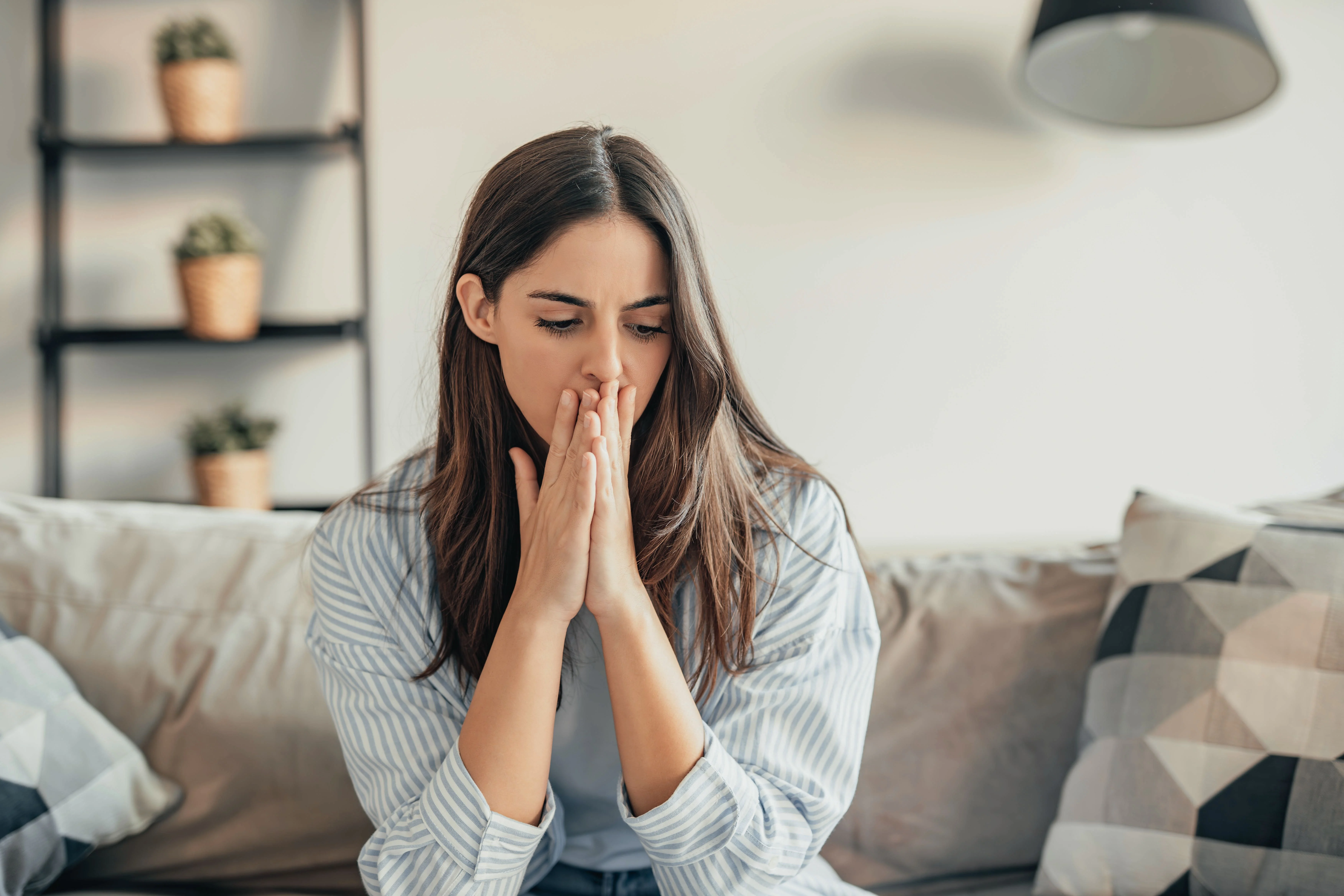 A worried young woman sits on a sofa, hands covering her mouth, with shelves of plants in the background