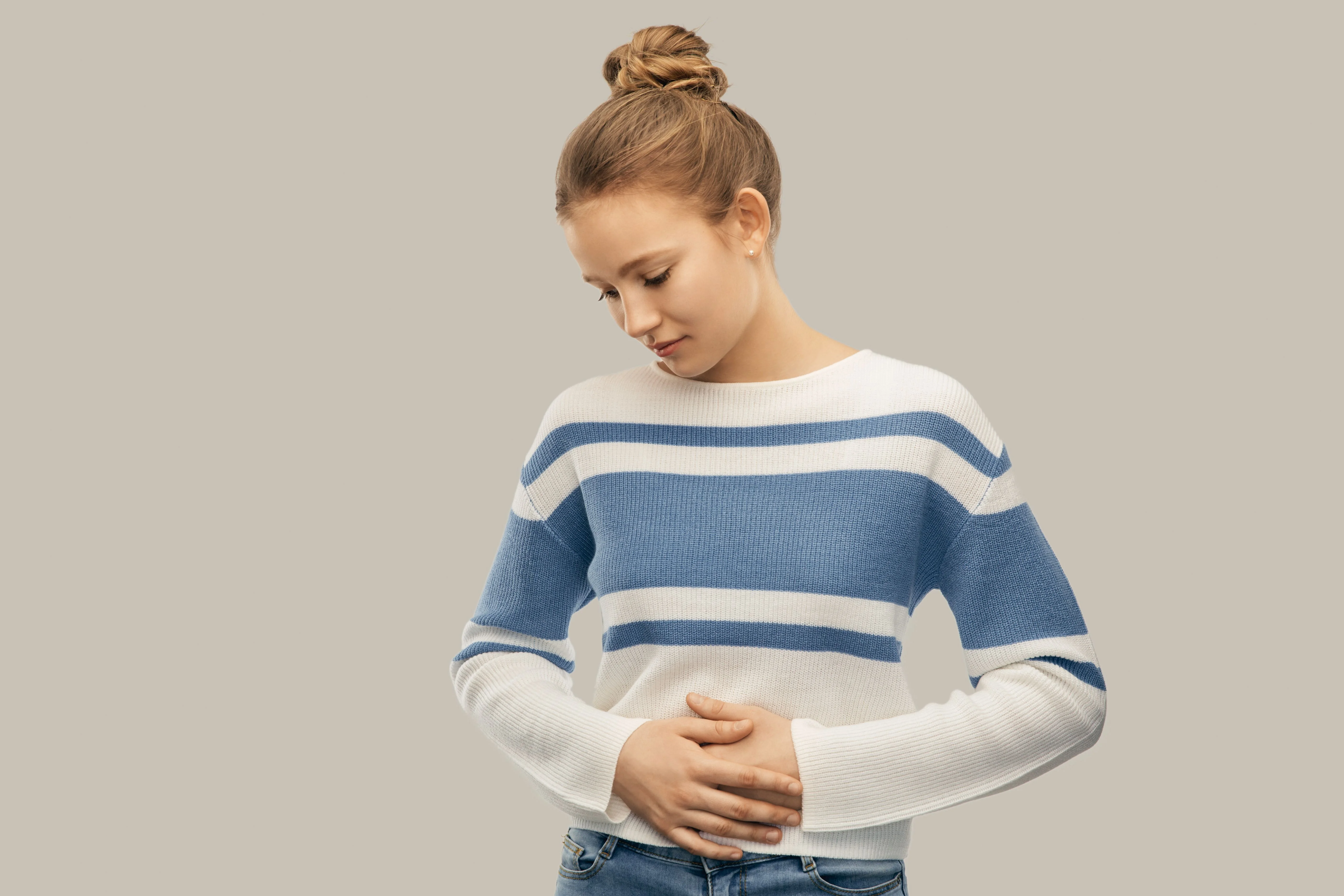 Mujer joven con un suéter a rayas azul y blanco, mirando hacia abajo y sosteniendo su abdomen, sobre un fondo neutro