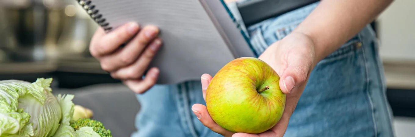 A person holds a green apple in one hand while holding a notebook in the other, surrounded by fresh vegetables