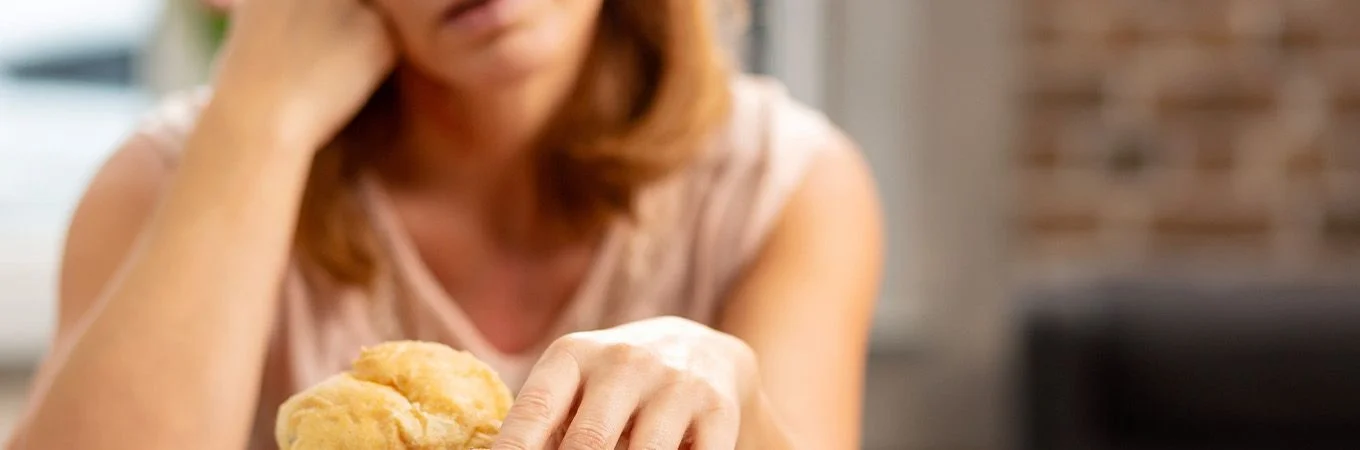 A woman looks pensive while resting her face on her hand, staring at a bun on the table in front of her