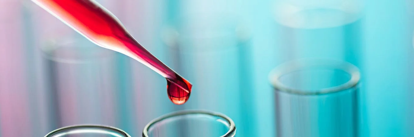 Eine Pipette lässt einen Tropfen roter Flüssigkeit in ein klares Reagenzglas fallen, vor blauem Hintergrund