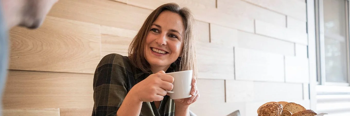 A smiling woman holding a coffee cup in a cozy setting with light wooden walls and a plate of pastries nearby