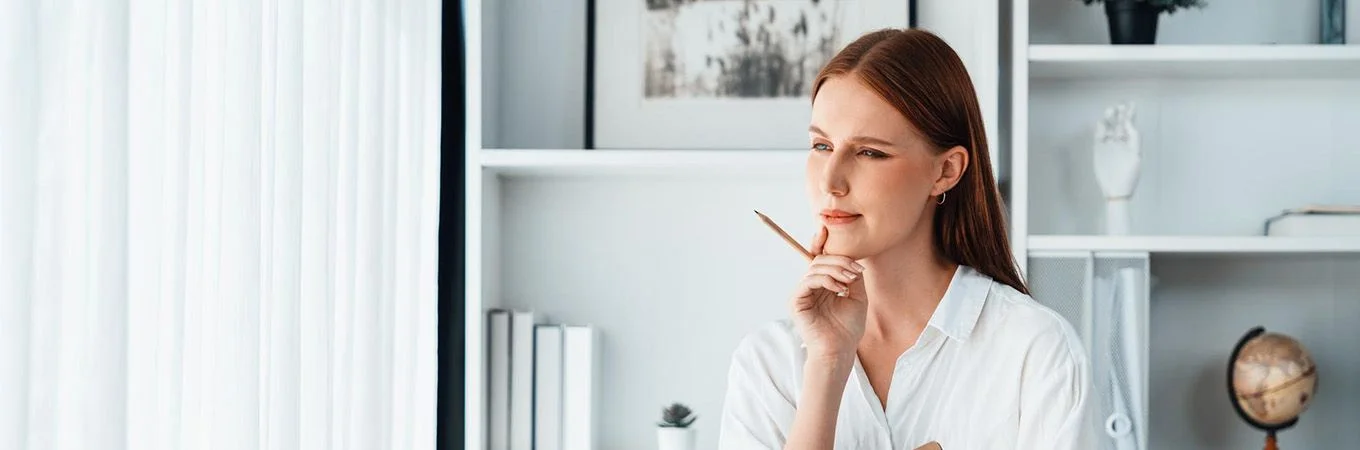 Young woman in a bright room thoughtfully holding a pencil, with shelves and art in the background