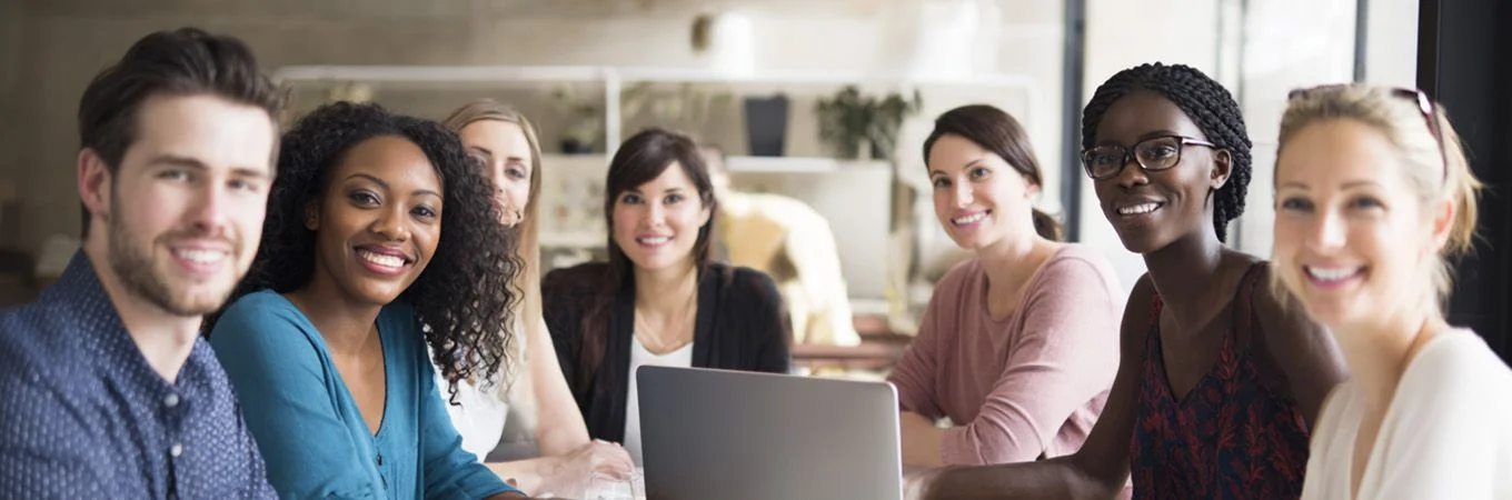 Eine vielfältige Gruppe von sieben Personen lächelt an einem Tisch mit einem Laptop in einem modernen Büro