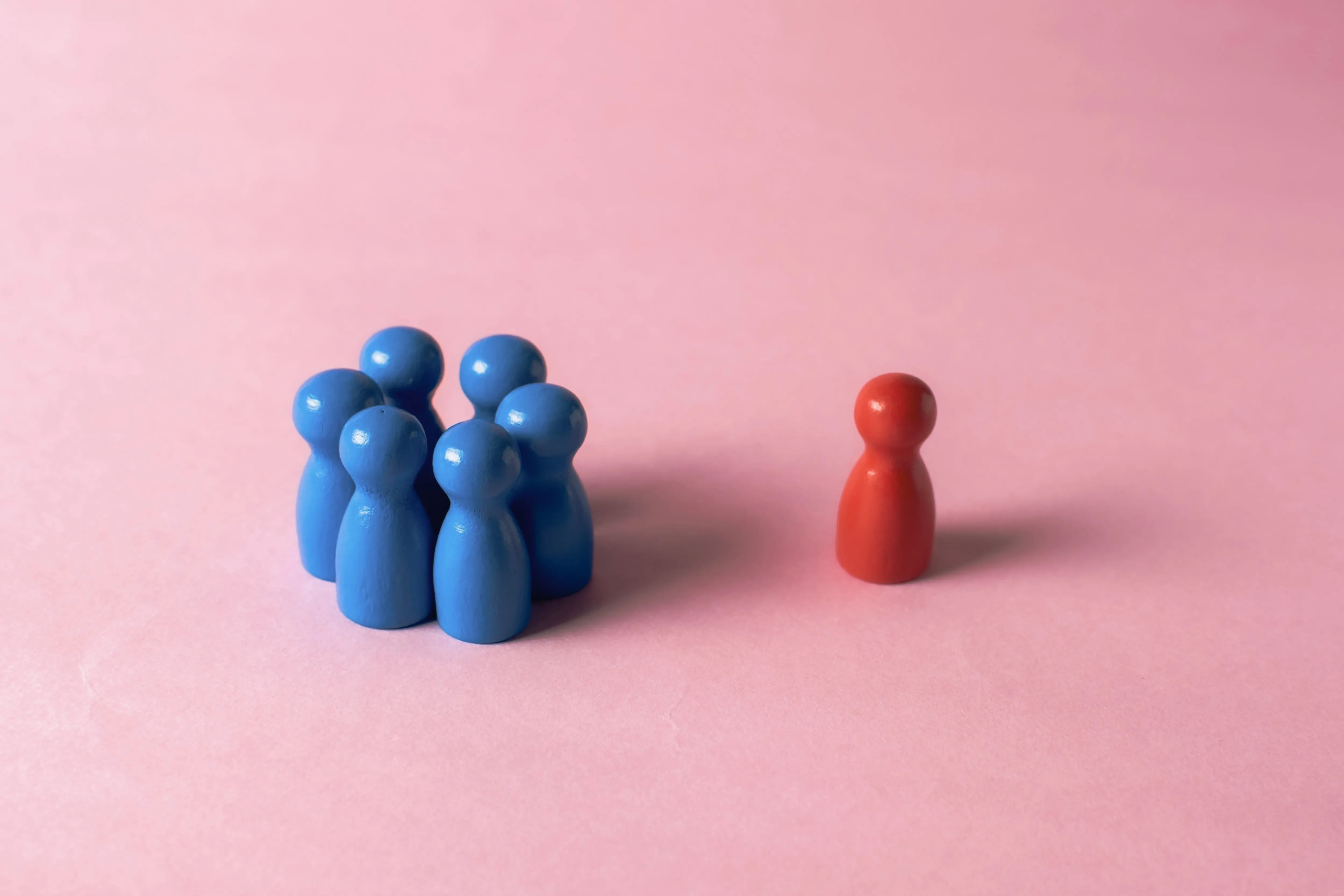 Six blue figurines huddle together against a pink background, while a single red figurine stands apart
