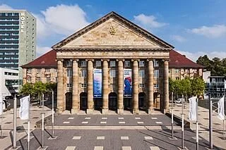 Historisches Gebäude mit fünf Säulen, mit Fahnen, umgeben von moderner Architektur und Bäumen unter blauem Himmel