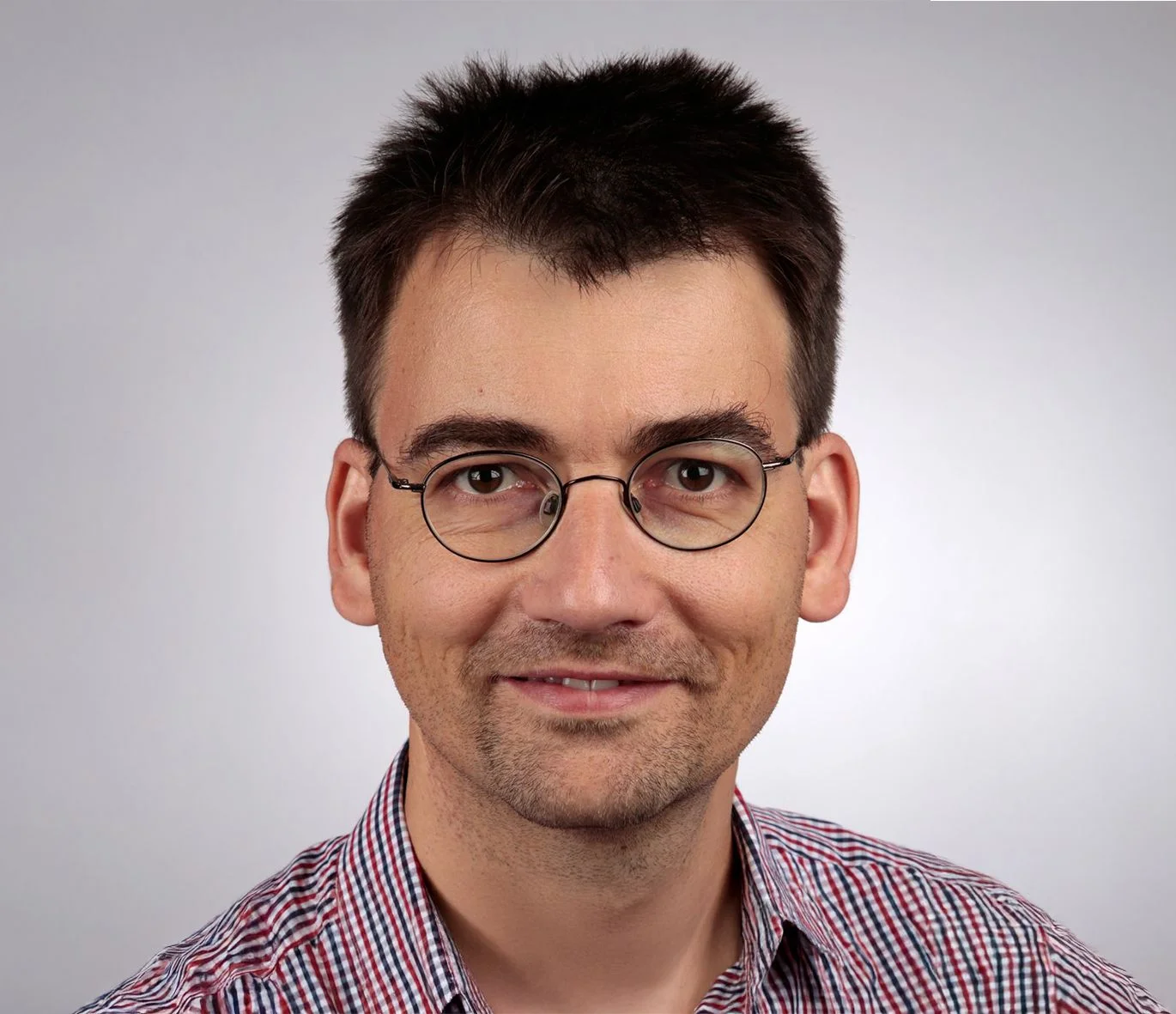 Uomo con capelli corti scuri e occhiali rotondi sorride, indossa una camicia a quadri su uno sfondo neutro