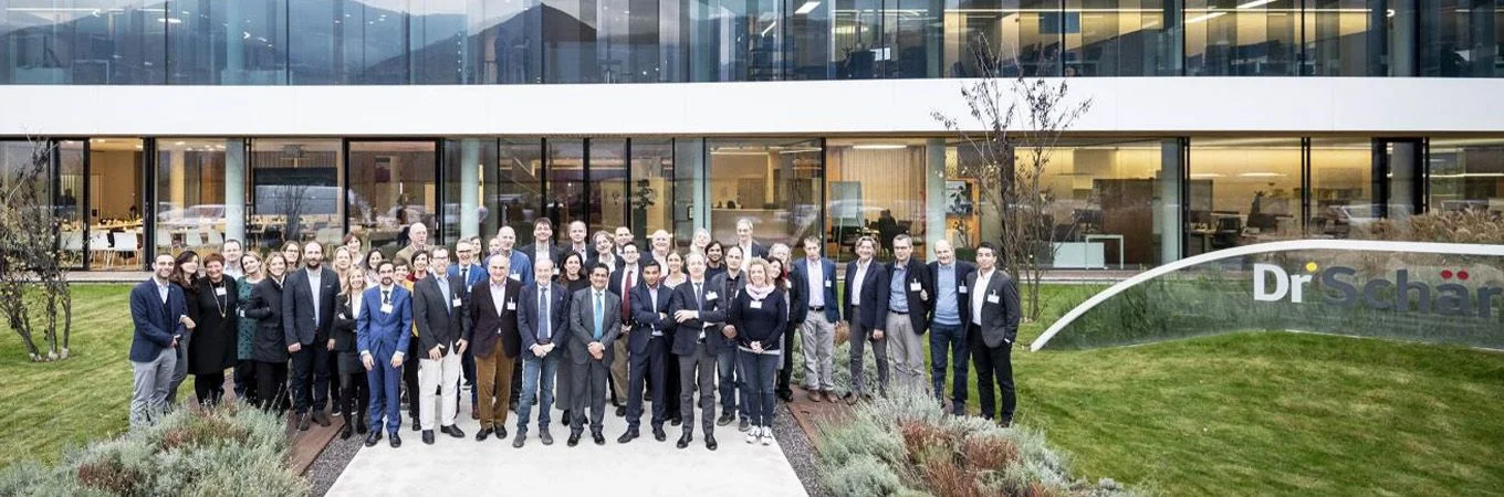 Un grupo diverso de profesionales posa frente a un edificio moderno con césped verde y el logo Dr. Schär