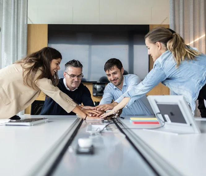 Four colleagues gather around a table, placing their hands together in the center, smiling and engaged