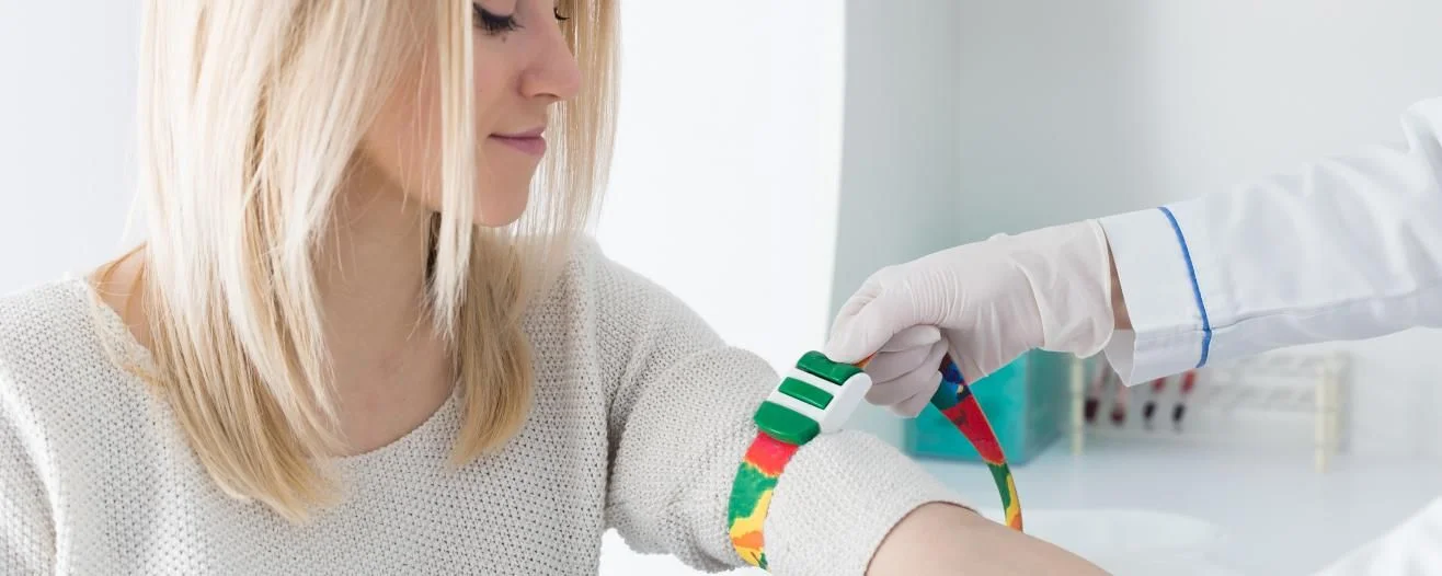 A woman with long blonde hair is having a colorful tourniquet applied to her arm by a gloved medical professional