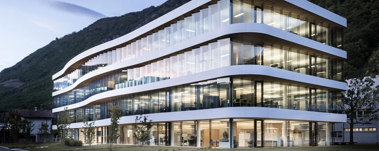 Edificio per uffici moderno con facciata in vetro curvato, circondato da verde e montagne sullo sfondo