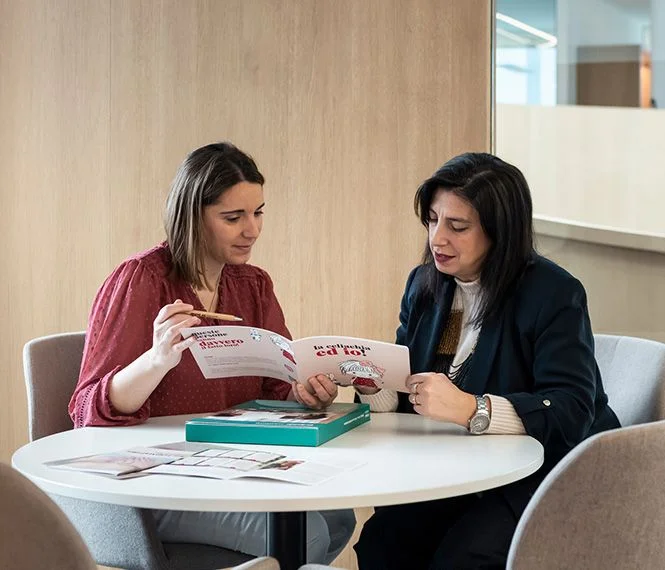 Two women examine brochures at a round table in a modern office space, engaged in discussion
