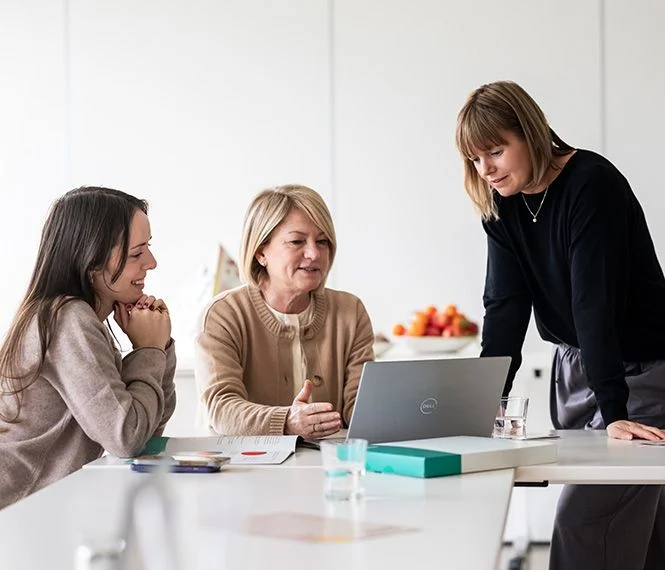 Three women engage in discussion over a laptop in a bright, modern office, with fruit in a bowl nearby