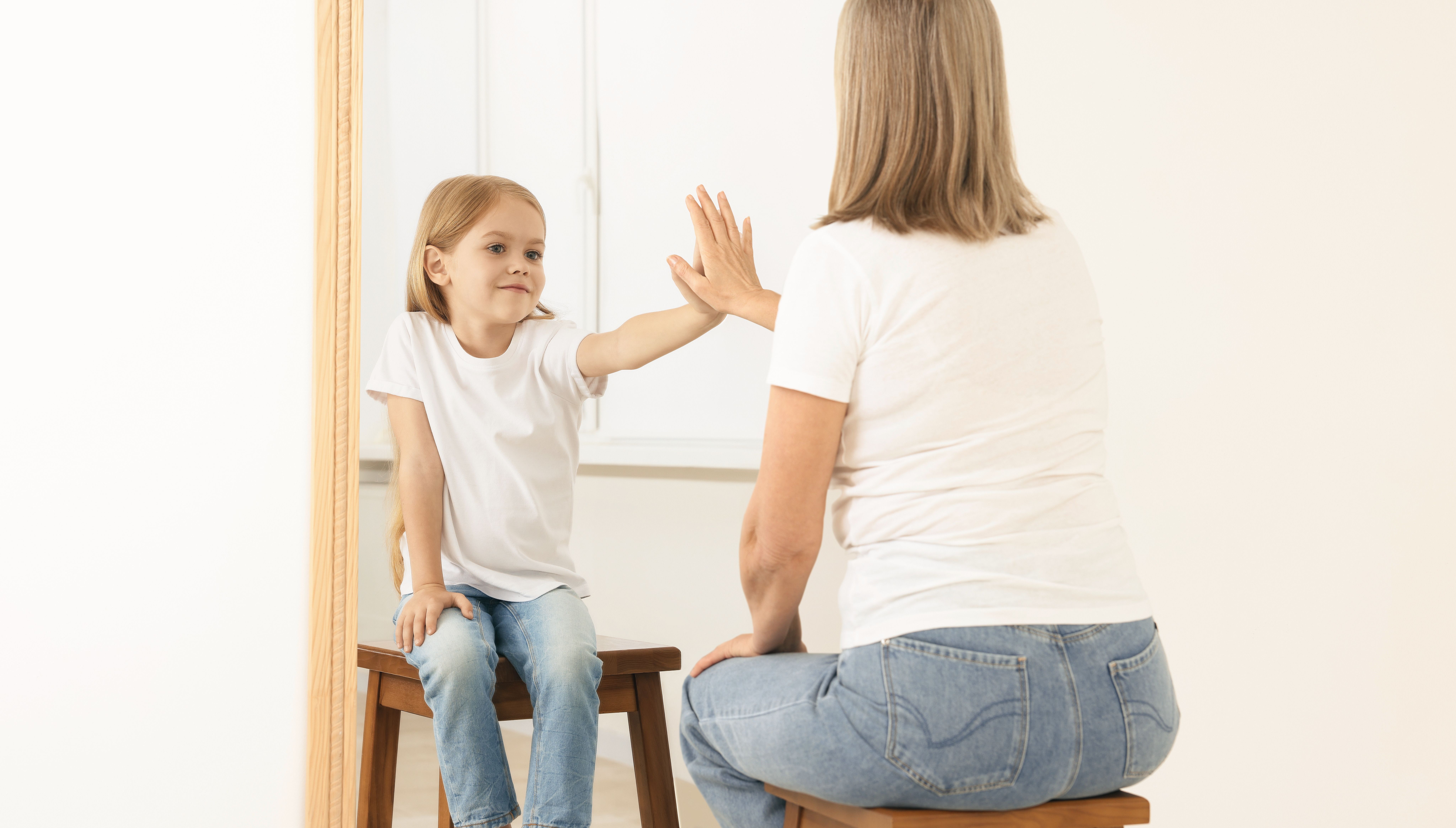 Una bambina e una donna sedute su sgabelli, si allungano l'una verso l'altra attraverso uno specchio in un momento gioioso